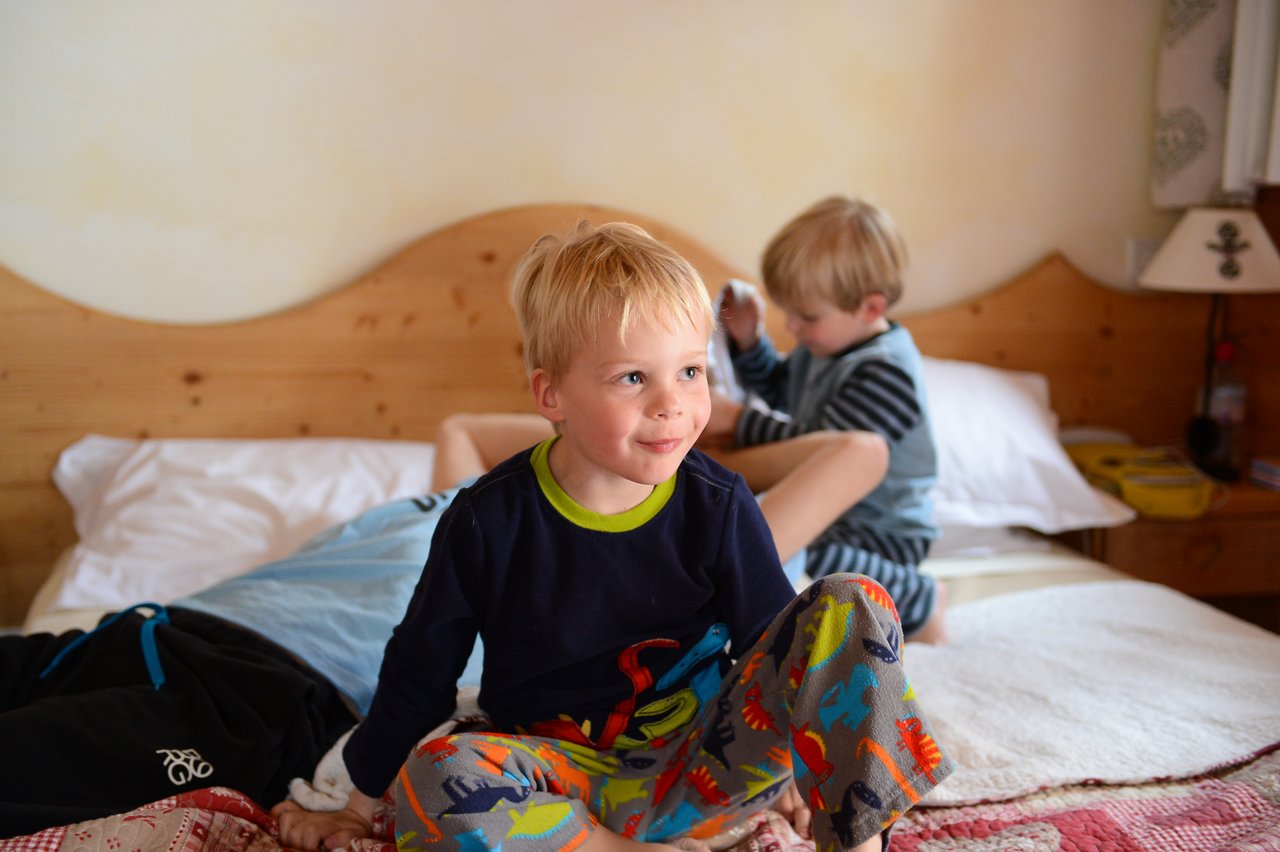 A young child sits on a bed, smiling, while another child plays in the background.