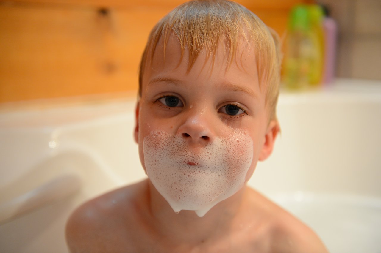A young child in a bathtub with soap bubbles on their face, resembling a beard.