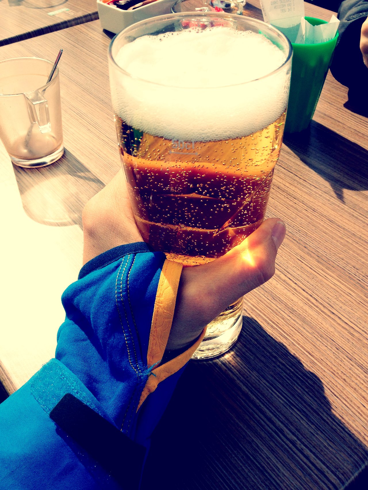 A person in a blue jacket holds a glass of beer with foam on top at a wooden table.