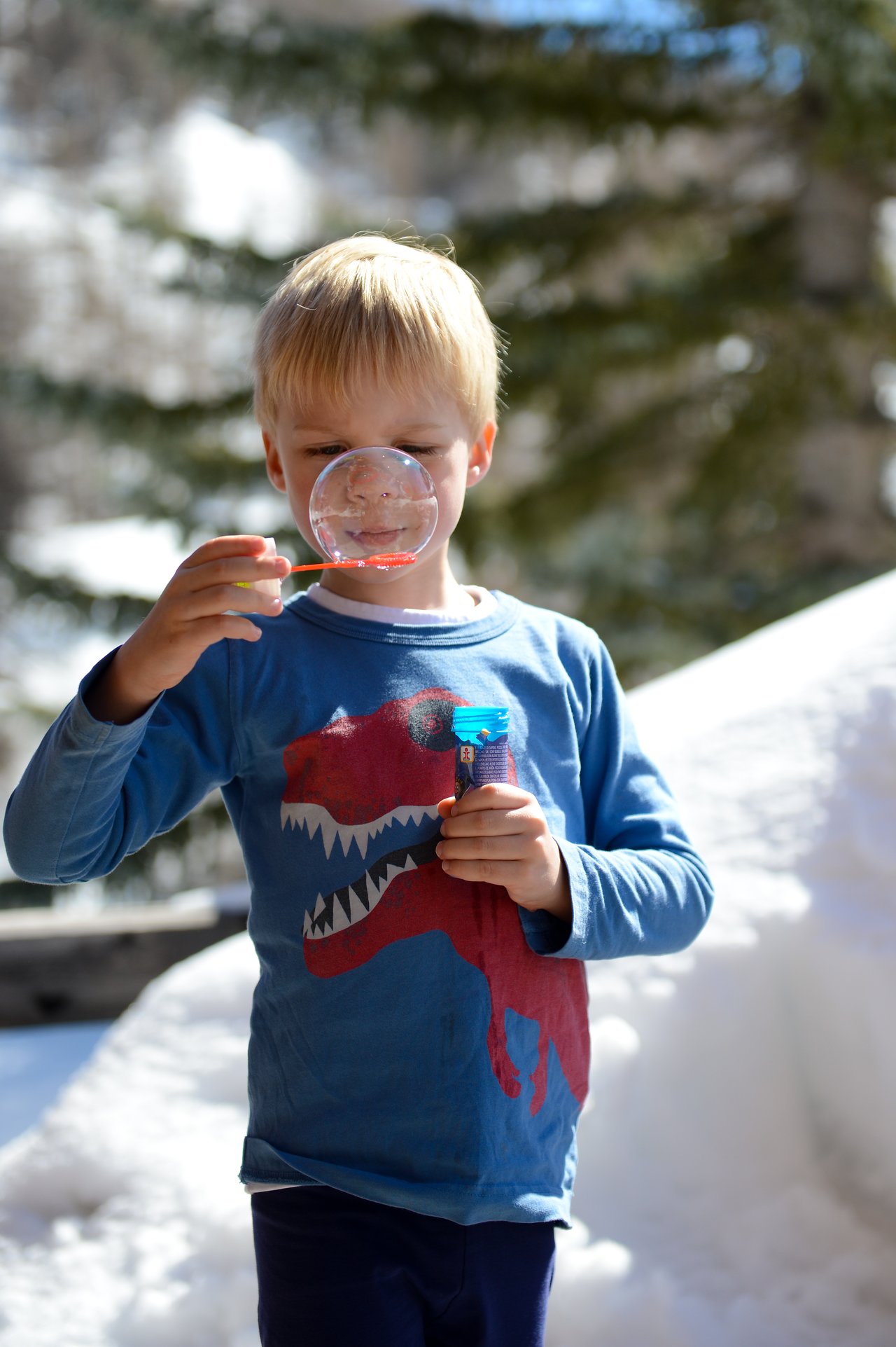 A young child in a blue shirt blows a bubble using a wand while holding a bottle of bubble solution.