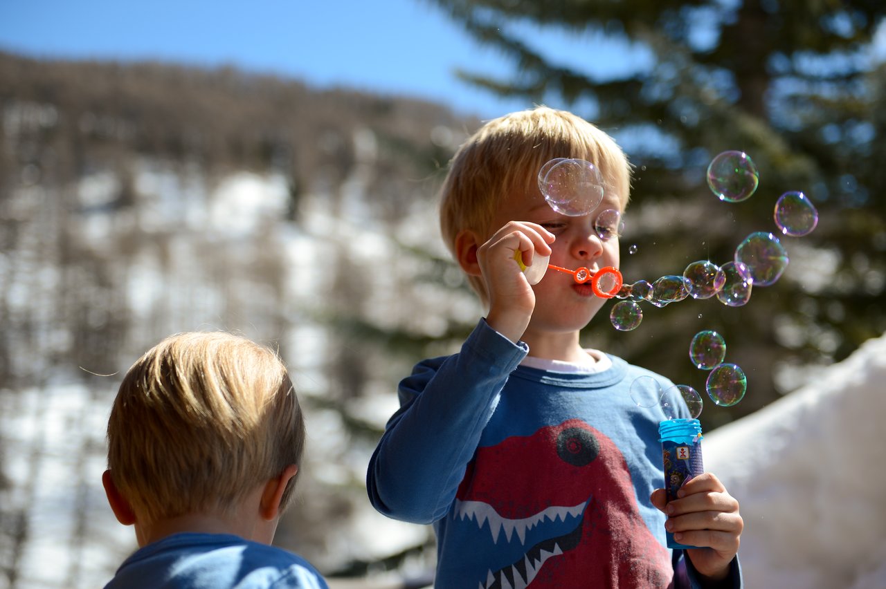 A child in a blue shirt blows bubbles while another child watches from the foreground.