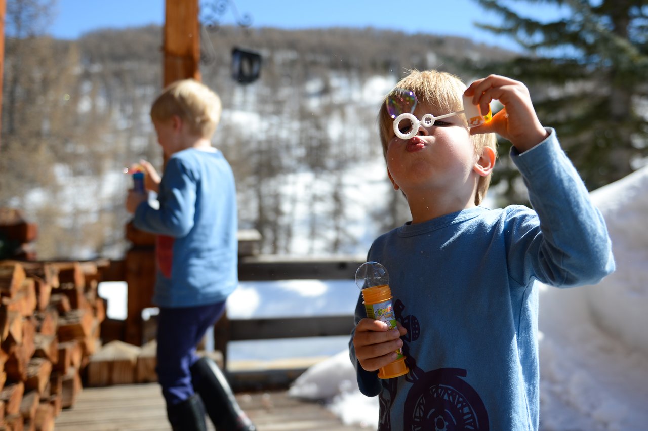 A child in a blue shirt blows bubbles while holding a bubble wand and container outdoors in a snowy setting.