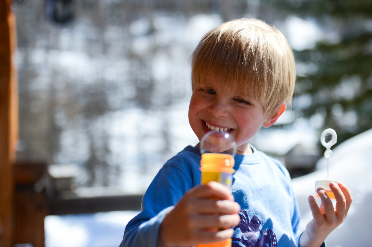 A smiling child in a blue shirt holds a bubble wand and bottle while blowing a bubble.