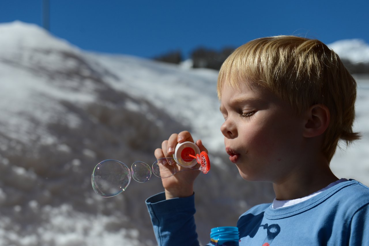 A child in a blue shirt blows soap bubbles using a red bubble wand outdoors.