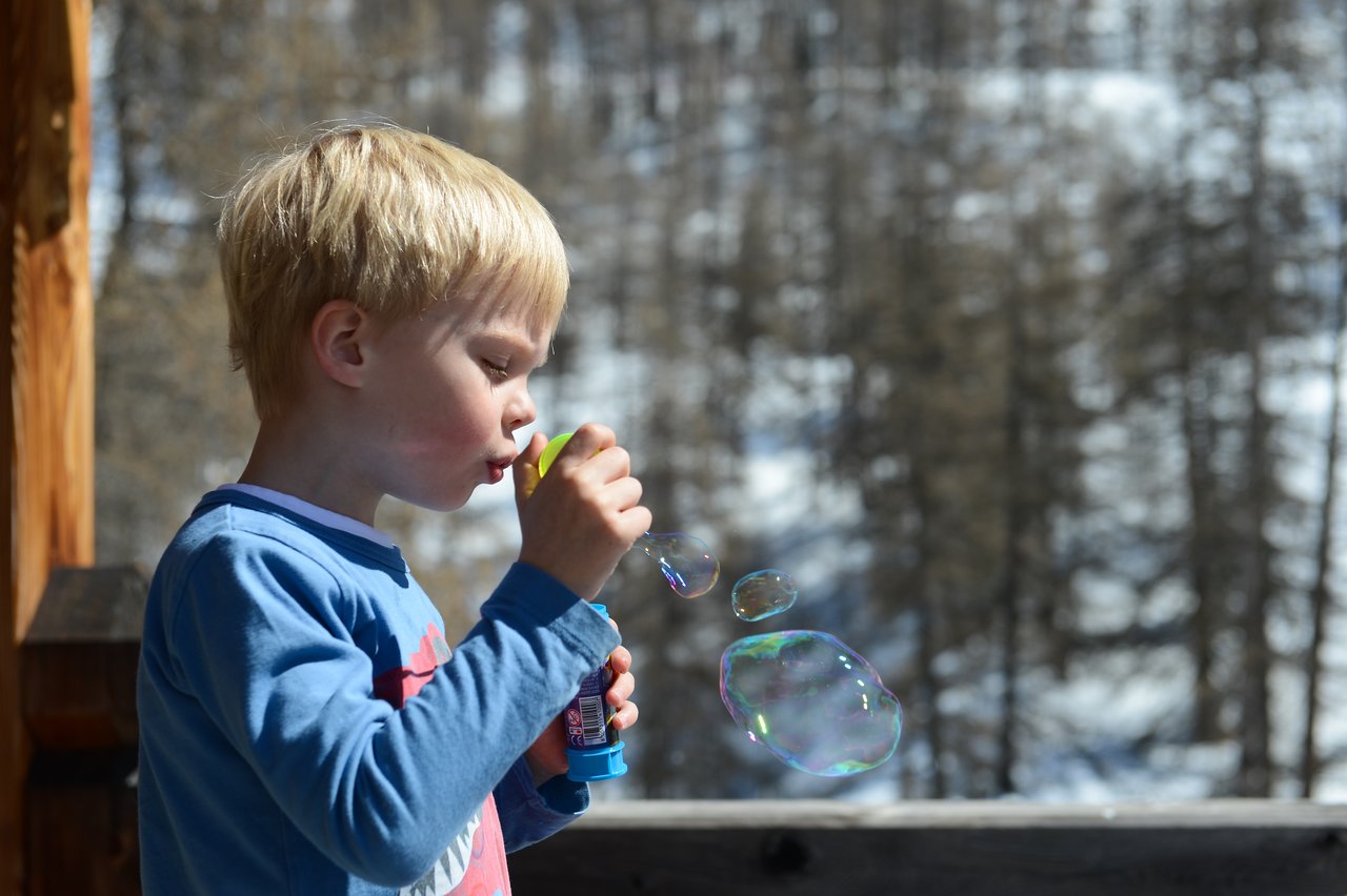 A child in a blue shirt blows soap bubbles while holding a bubble wand and container.