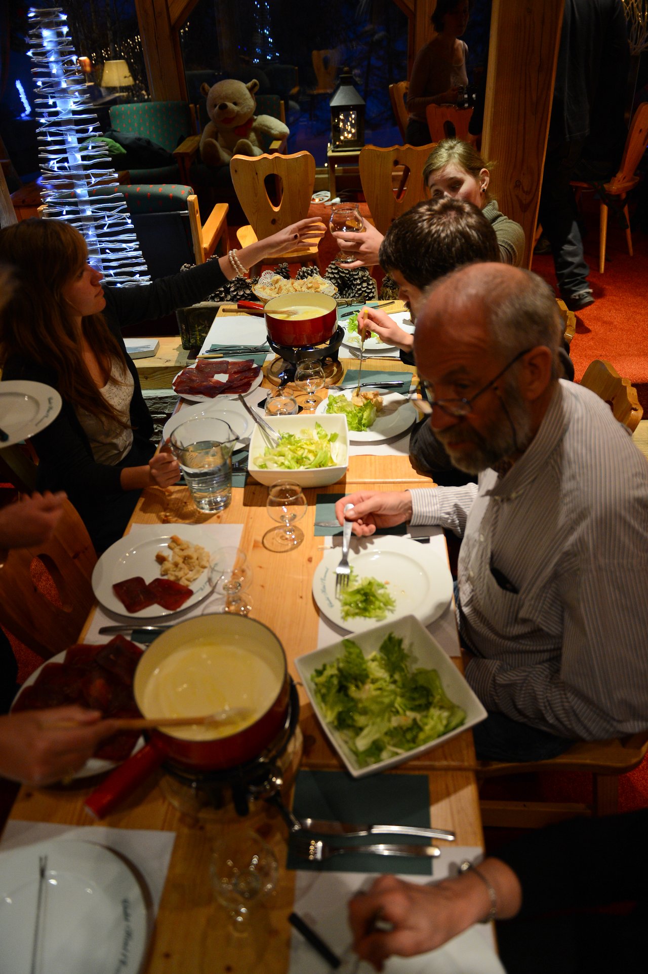 A group of people share a cheese fondue meal, dipping food and toasting drinks around a wooden table.