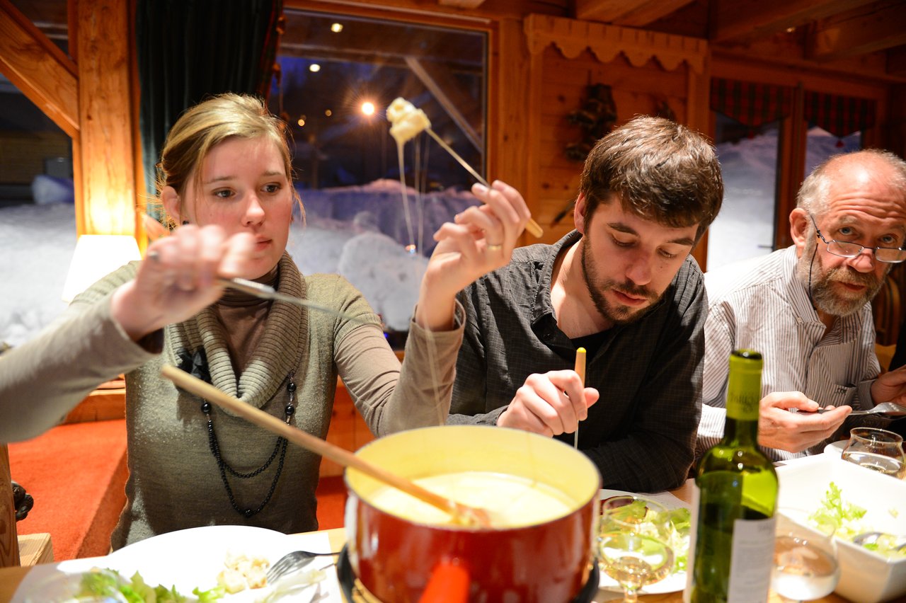 Three people enjoying a cheese fondue meal, dipping bread into a pot of melted cheese with skewers.