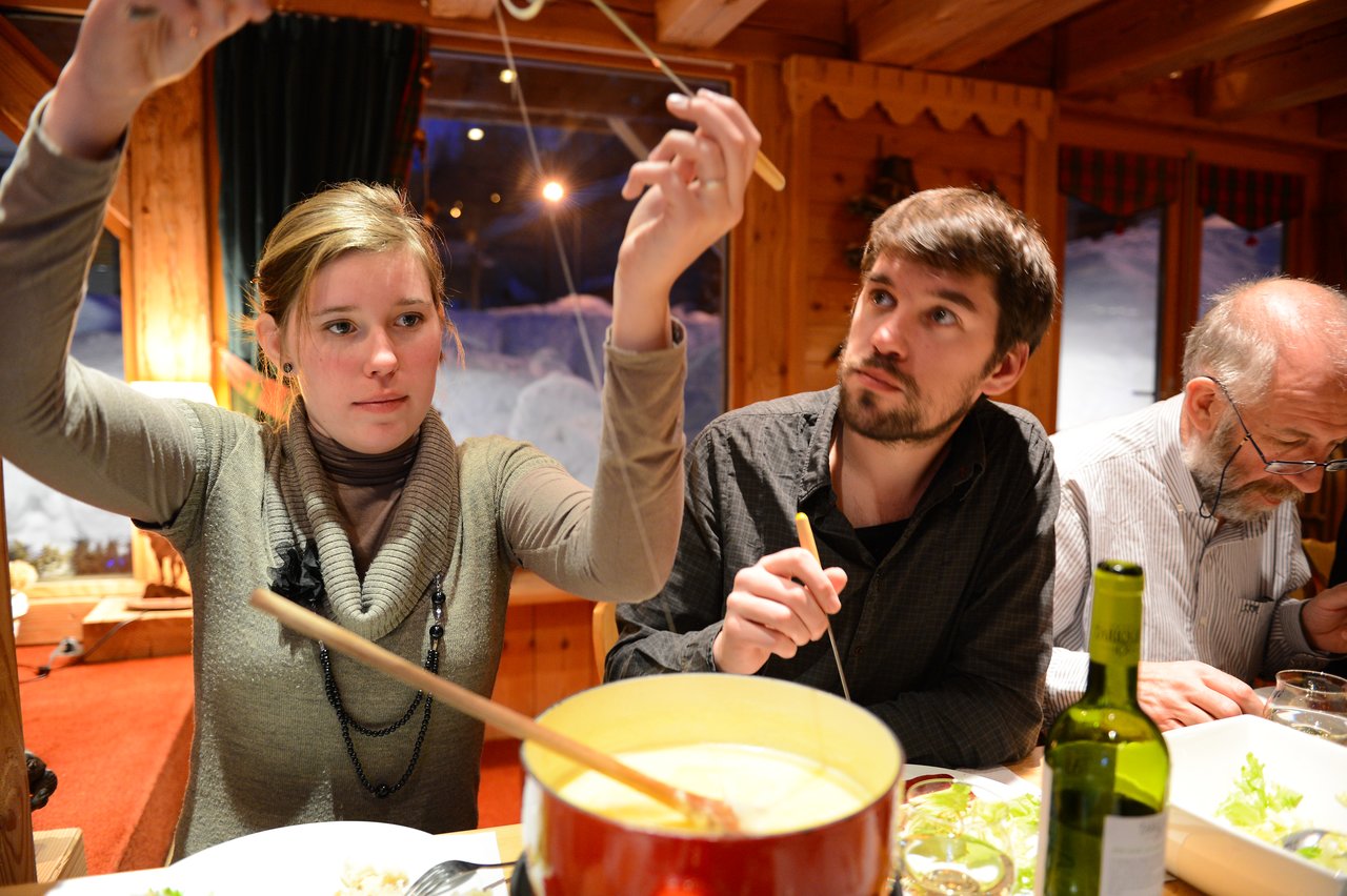 A woman lifts melted cheese with a fork while others sit around a table enjoying a fondue meal.