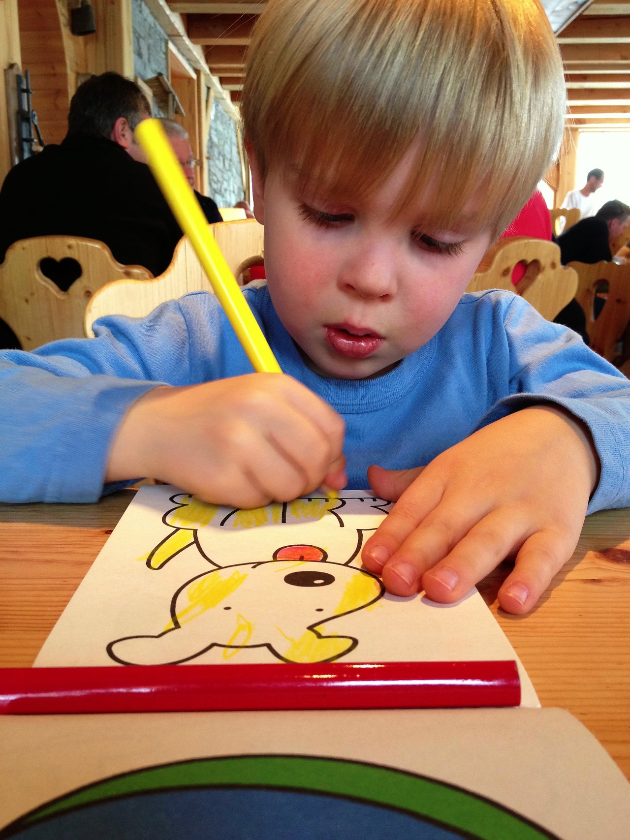 A young child in a blue shirt colors a picture of a dog with a yellow pencil.