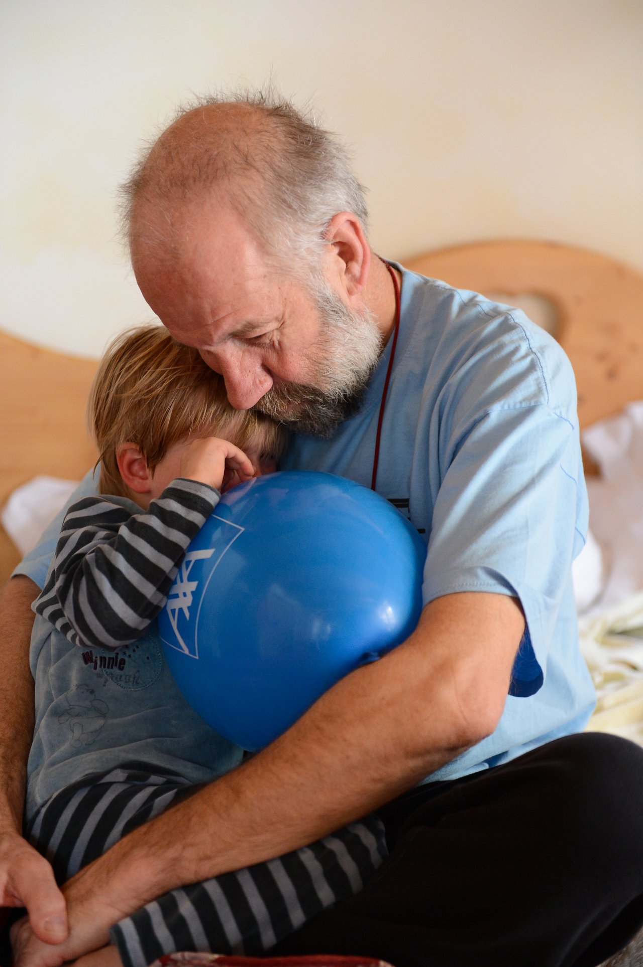 An older man hugs a crying child holding a blue balloon, comforting them while sitting on a bed.