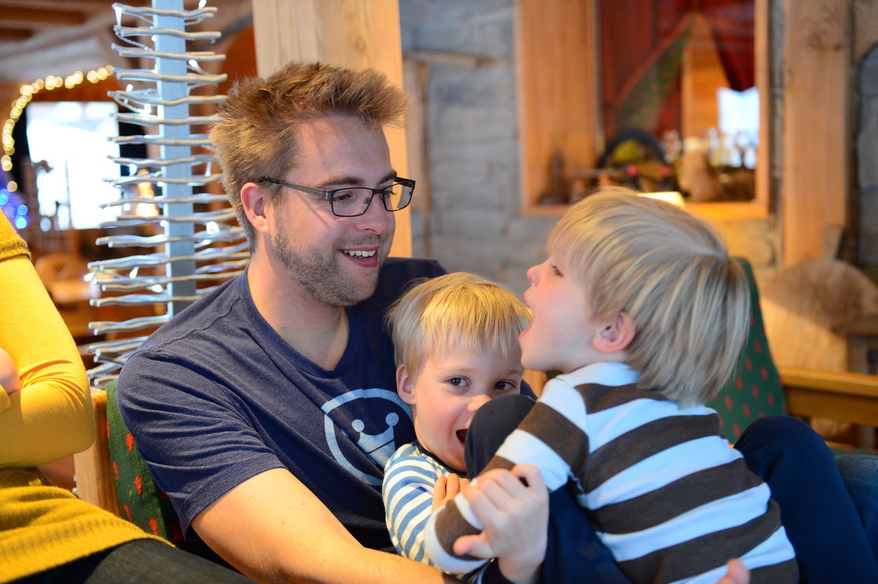 A father sits with his two young children, smiling as they play and interact closely with each other.