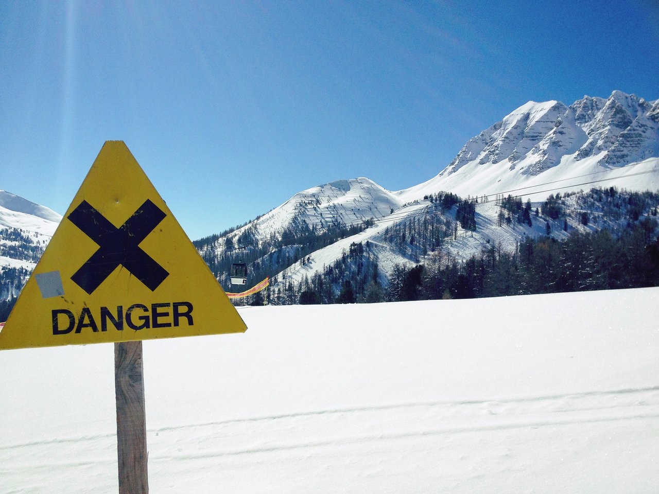 A yellow danger sign with a black X stands in a snowy mountain landscape under a clear blue sky.