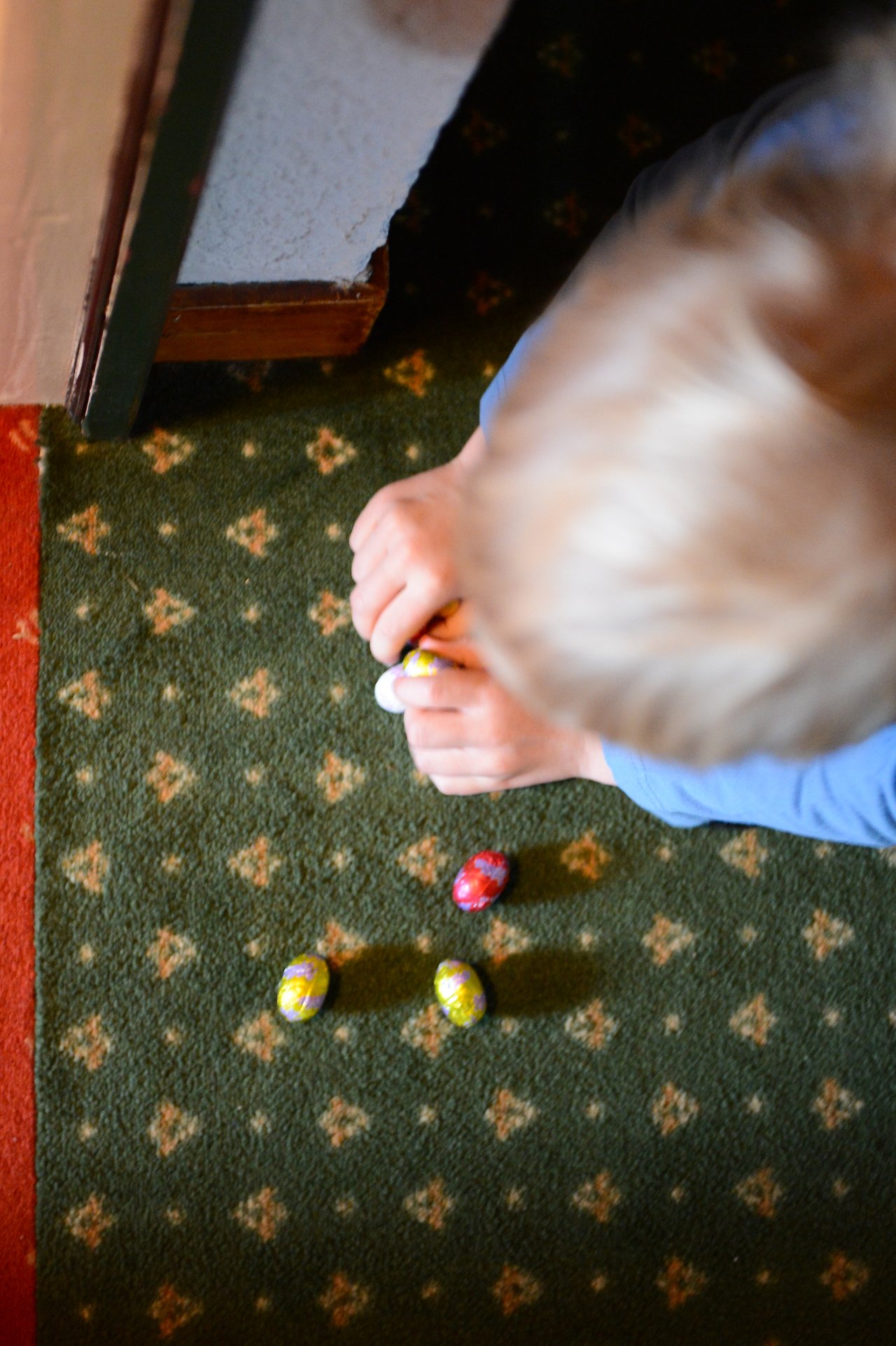 A child in a blue shirt picks up a small Easter egg from a green patterned carpet.