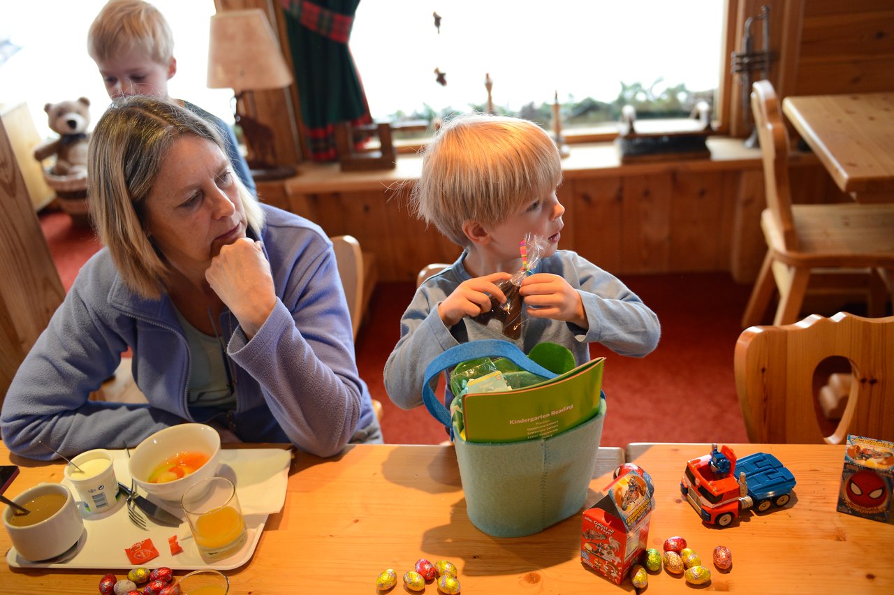 A young child opens Easter candy while sitting at a table with a woman and festive treats.