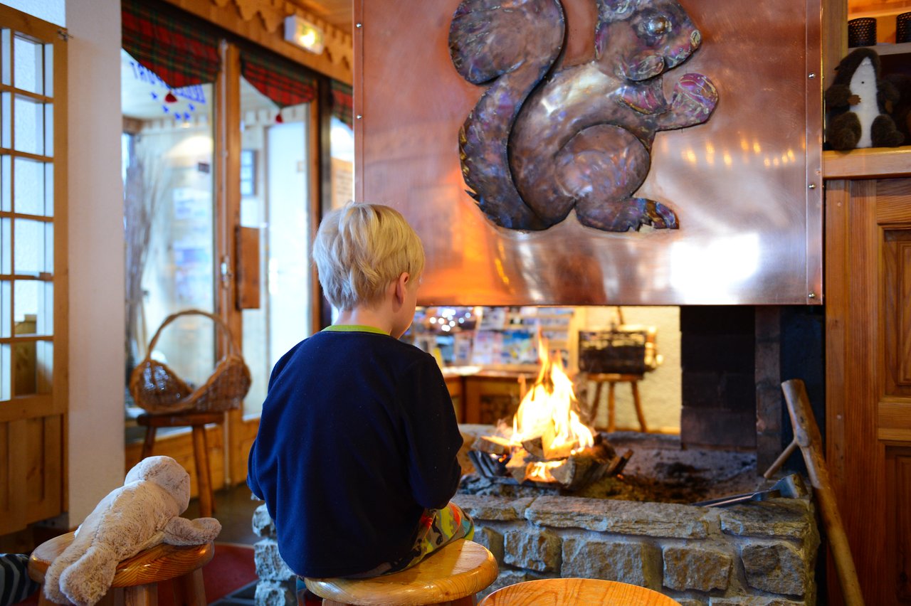 A child sits on a stool near a fireplace, facing the fire with a stuffed toy nearby.
