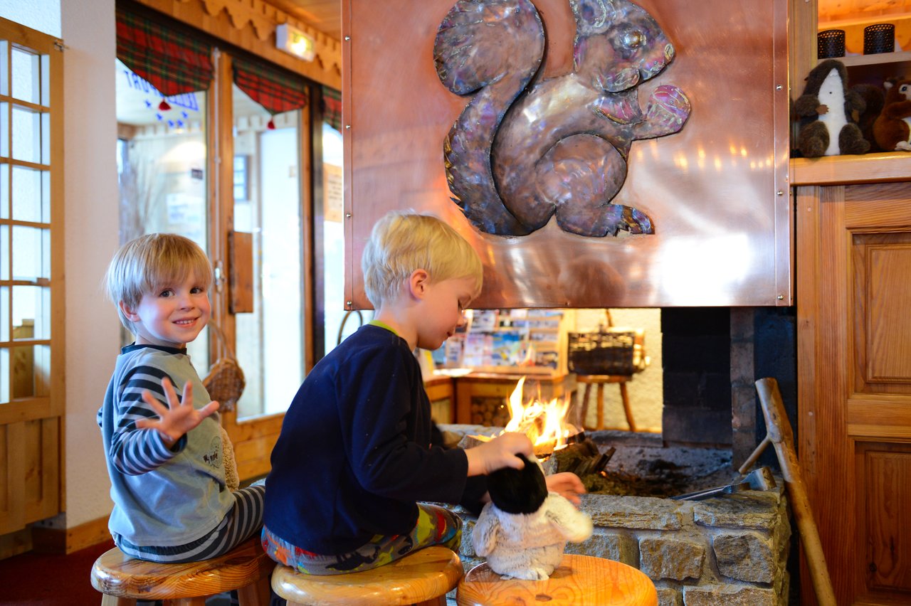 Two young children sit on stools near a fireplace, one waving and the other holding a stuffed animal.
