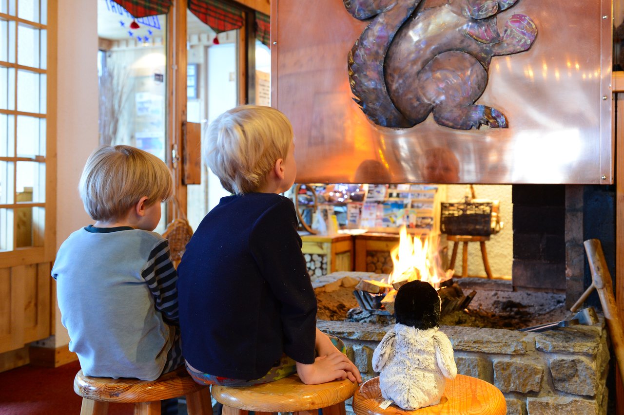 Two children sit on stools by a fireplace, watching the fire with a stuffed penguin toy beside them.