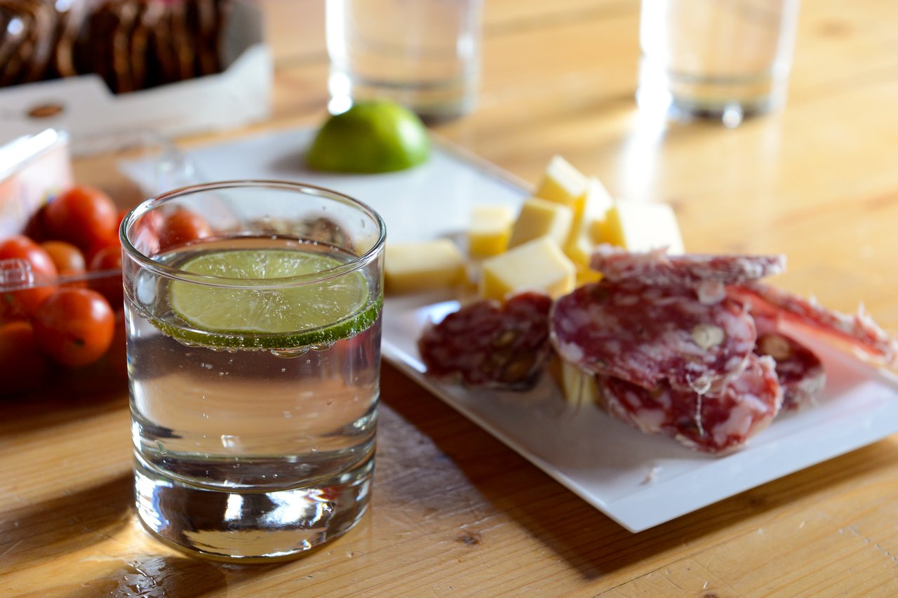 A glass of gin and tonic with a lime slice, next to a plate of cheese and salami.
