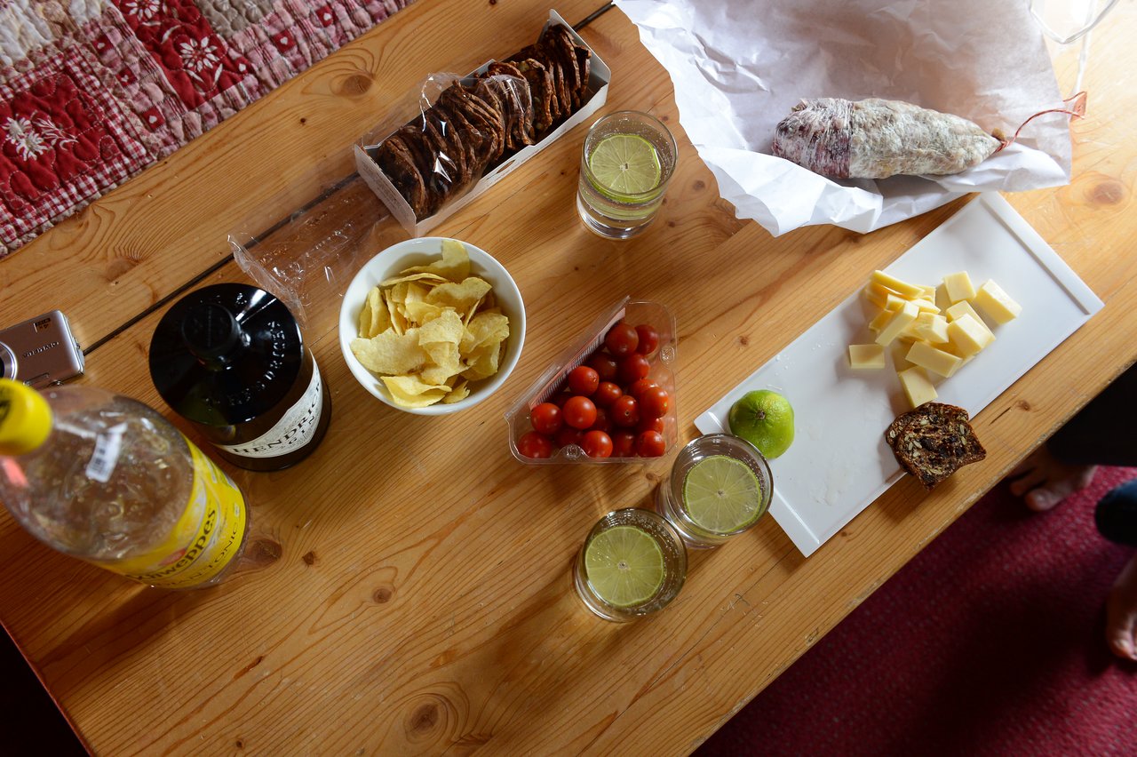 A wooden table with gin and tonic drinks, lime slices, cheese, crackers, chips, cherry tomatoes, and cured meat.