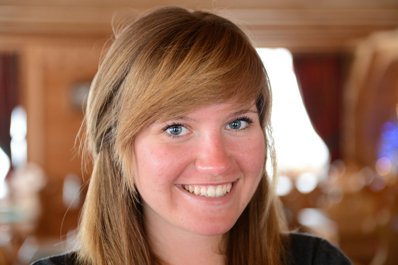 A woman with long brown hair smiles brightly, looking directly at the camera in a warmly lit indoor setting.
