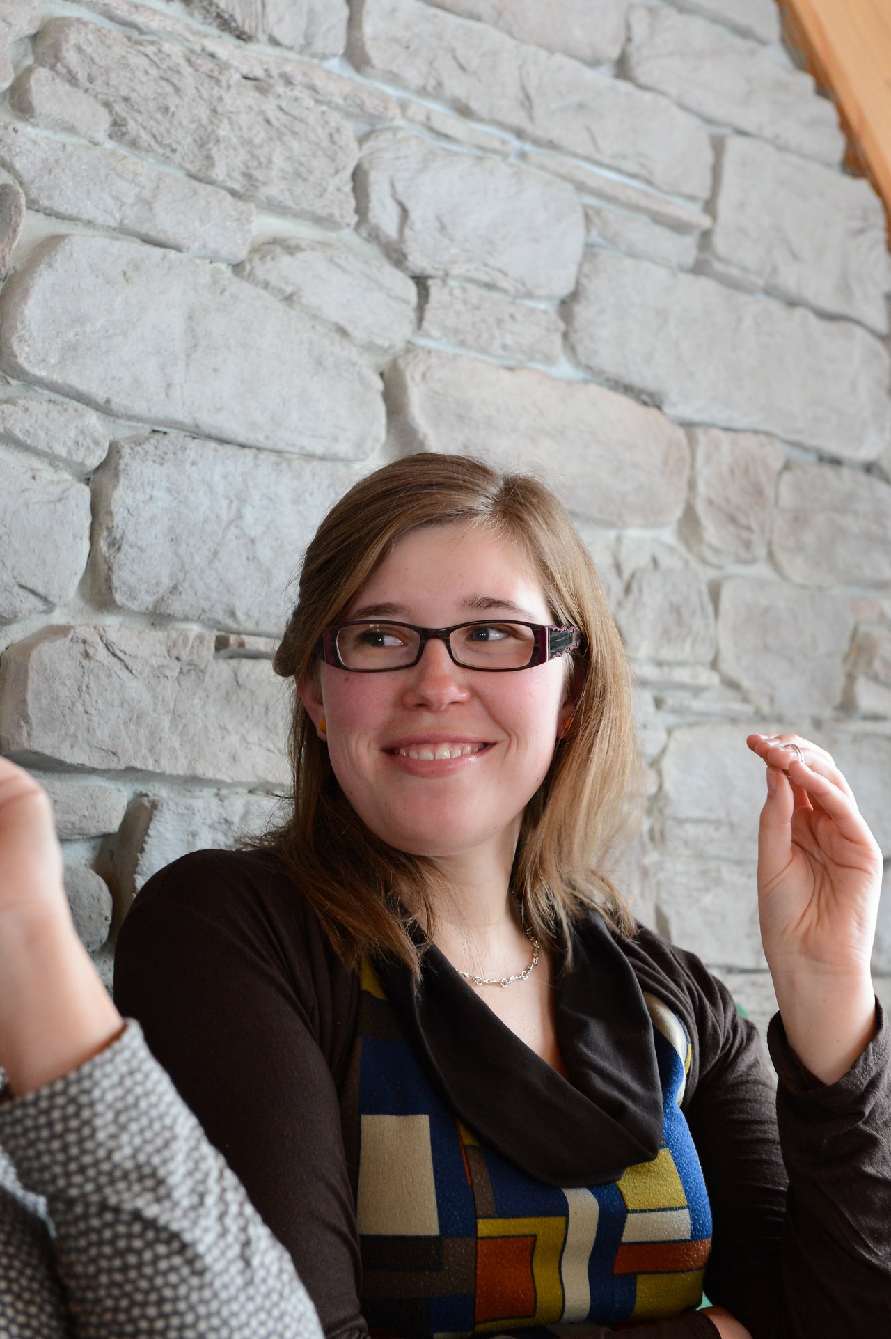 A woman with glasses smiles while gesturing with her hand, engaged in conversation with someone partially visible.
