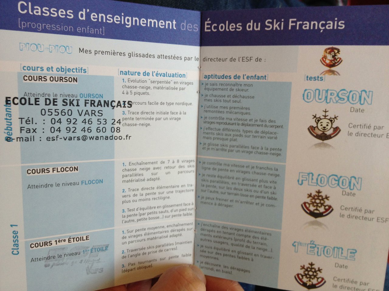 A person holds an open ski school booklet showing course levels, objectives, and certification tests for children's skiing progression.