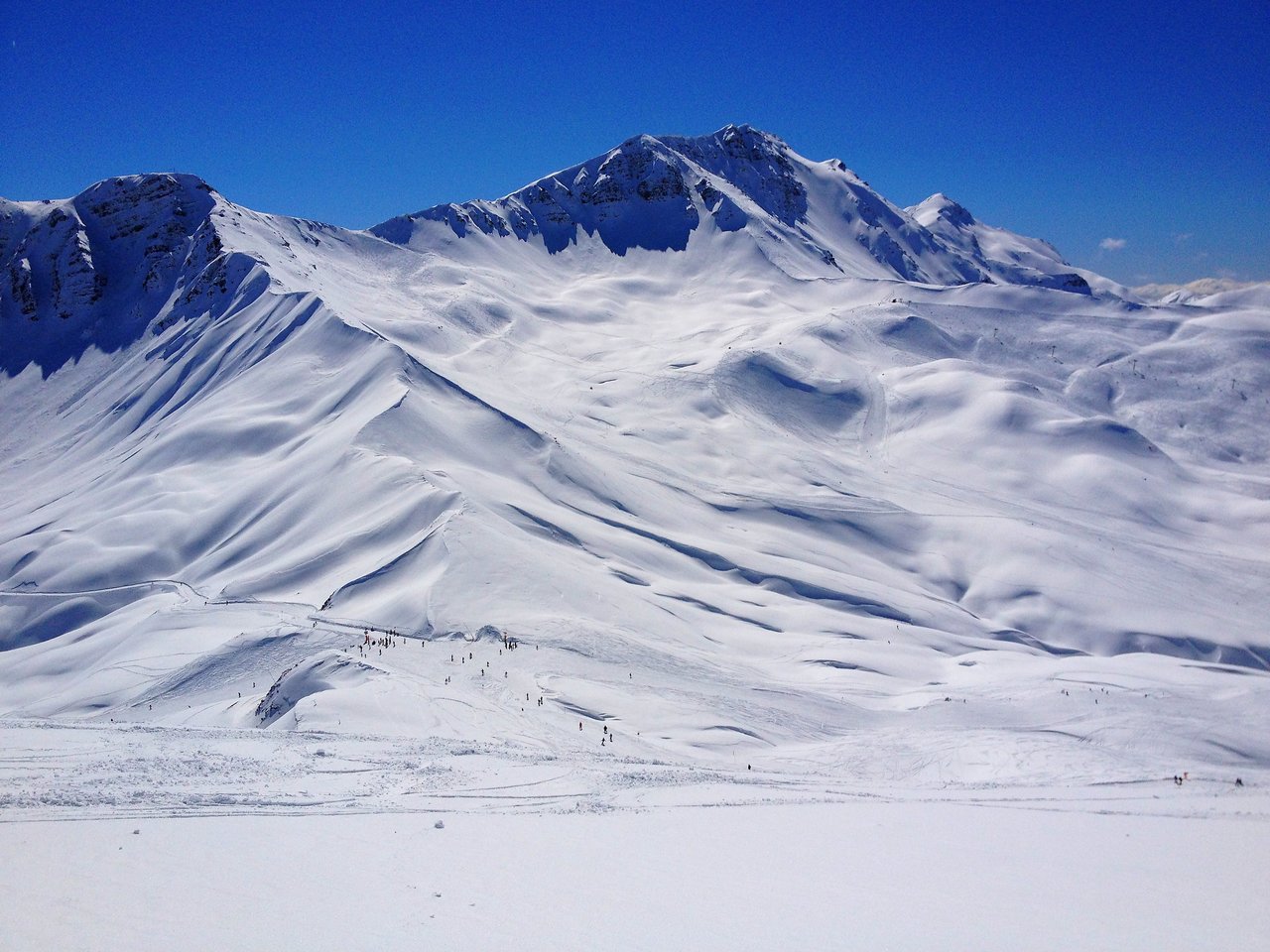 Snow-covered mountains with skiers scattered across the slopes under a clear blue sky.