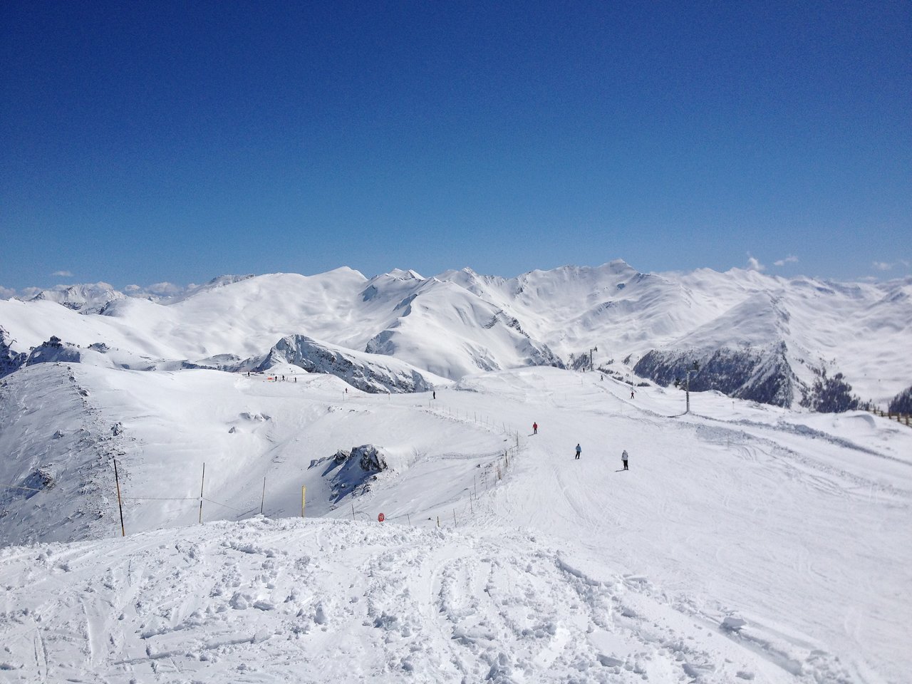 Skiers glide down a snowy mountain slope under a clear blue sky, surrounded by snow-covered peaks.