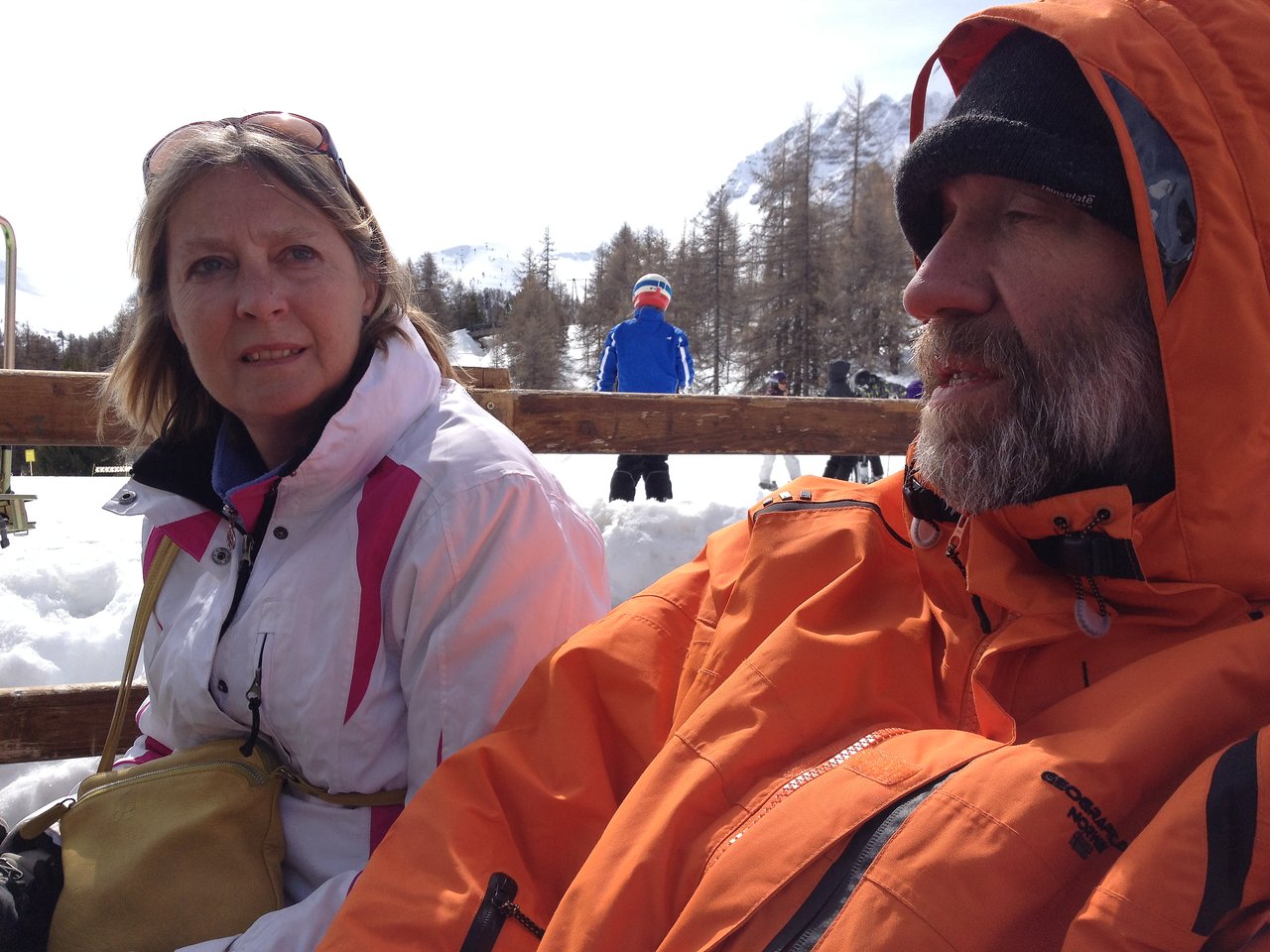 A woman and a bearded man in winter jackets sit on a bench in a snowy outdoor setting.