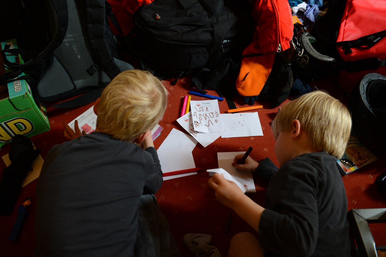 Two children lie on the floor, drawing on paper with markers, surrounded by backpacks and scattered art supplies.