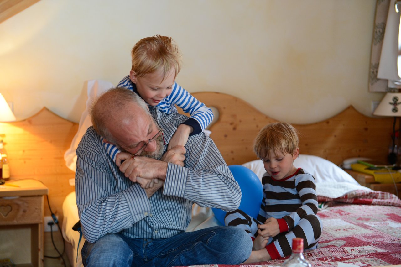 An older man sits on a bed, playfully hugging a young child while another child watches nearby.