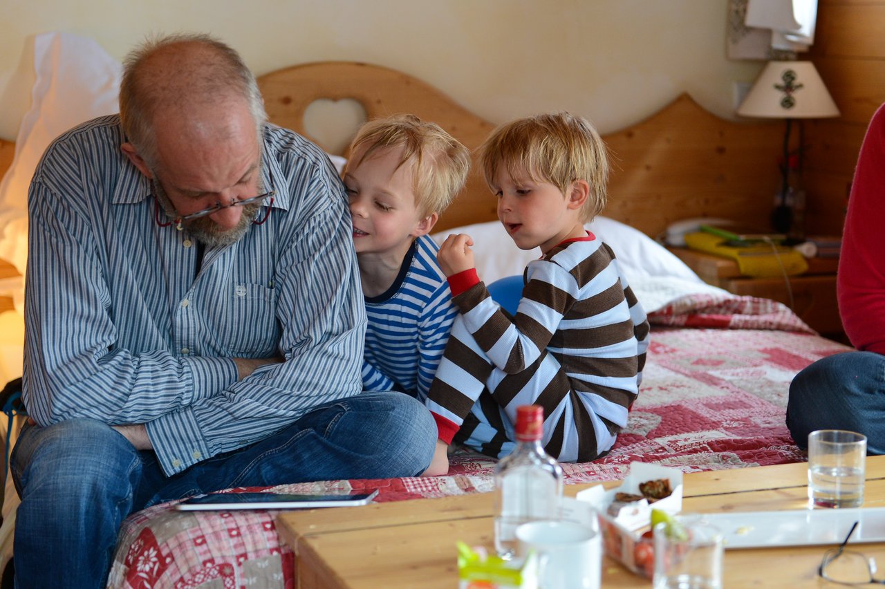 An older man sits on a bed reading a tablet while two young children in pajamas interact with him.