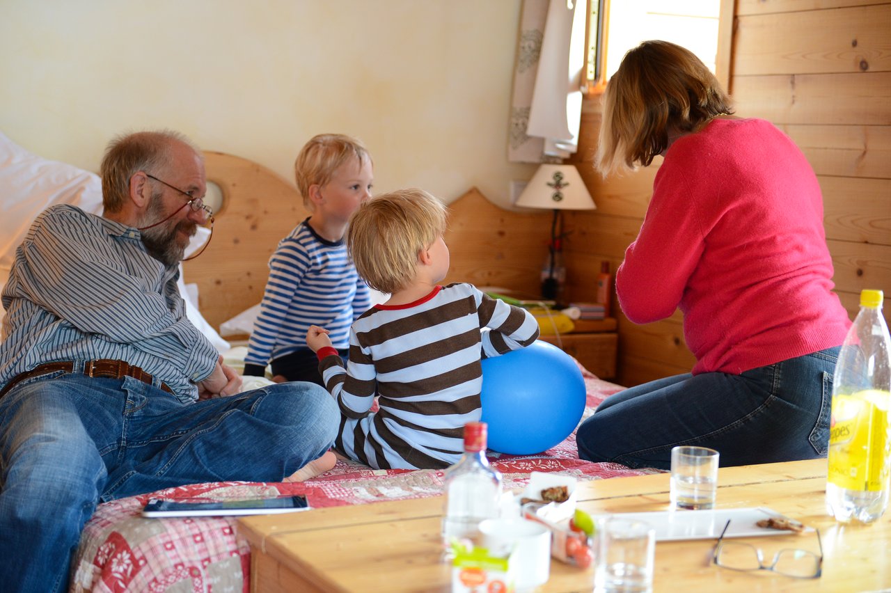 A man, two children, and a woman sit on a bed, interacting while one child holds a blue balloon.