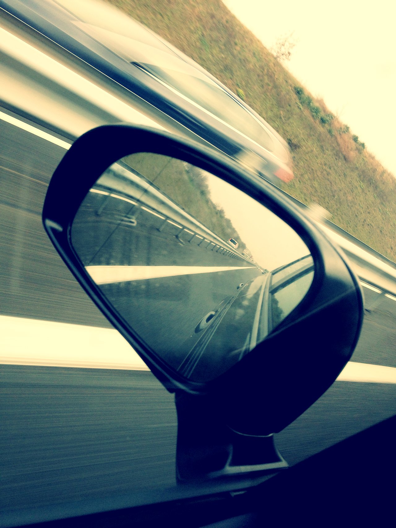 A car's side mirror reflects the road behind, while another vehicle passes by on the highway.