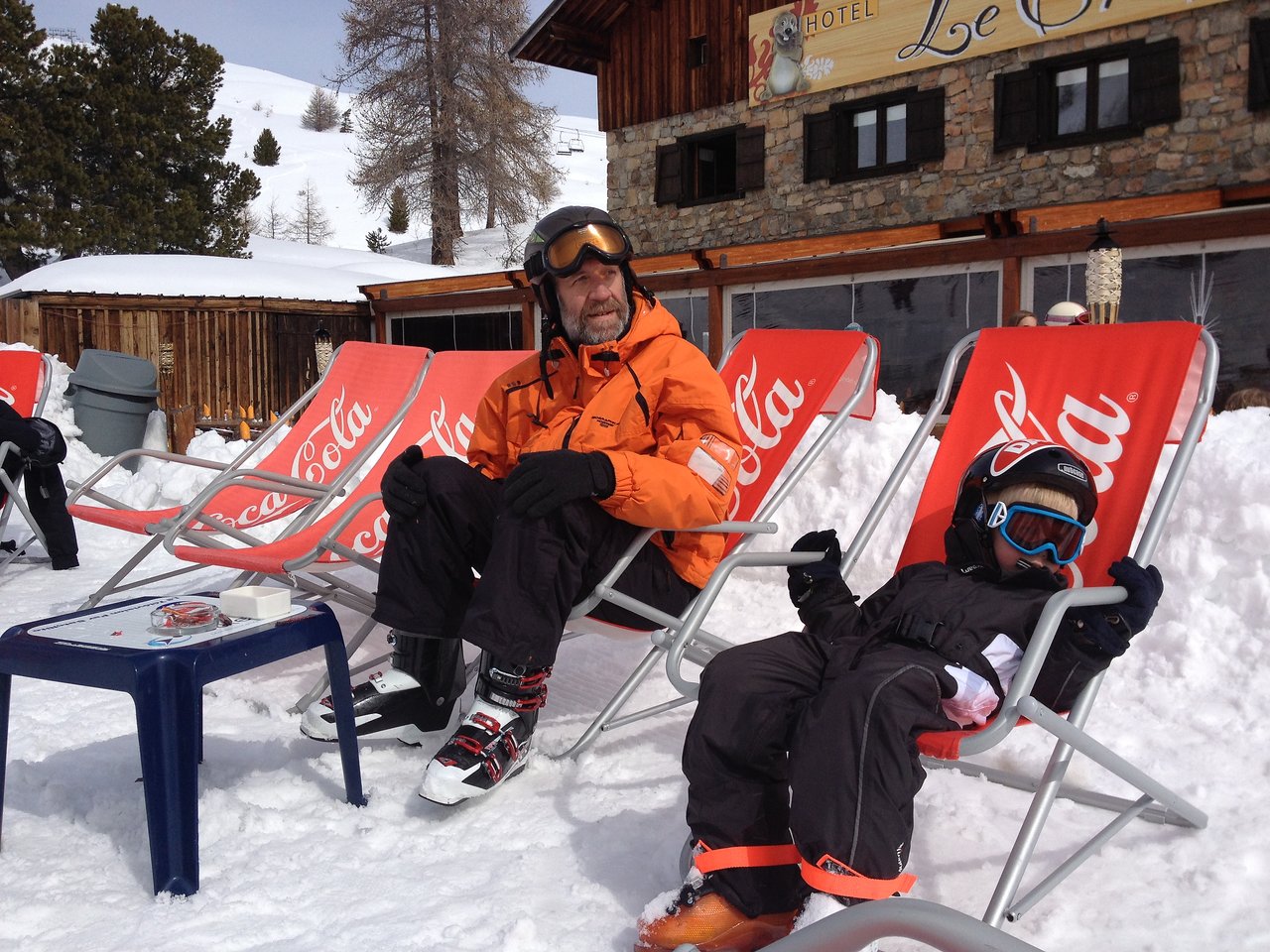 An older man and a child in ski gear relax on lounge chairs in the snow during a break.