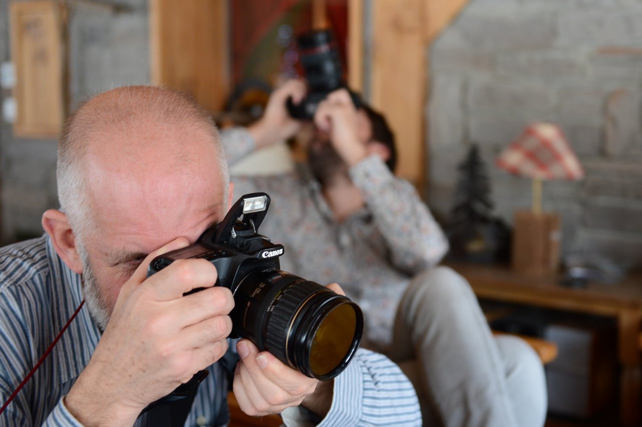 Two photographers holding cameras, one in the foreground focusing on a shot, the other in the background aiming upward.