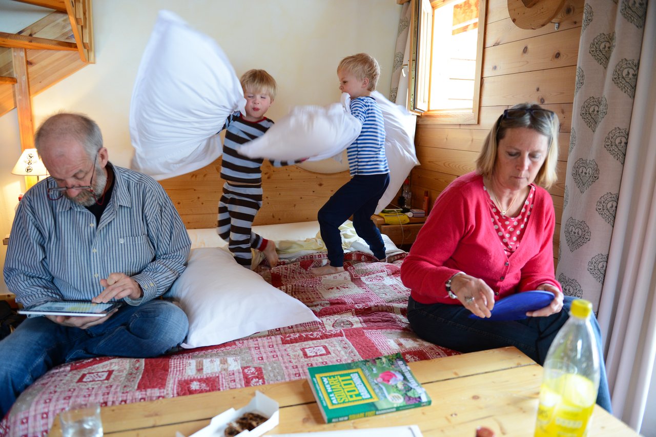 Two children on a bed have a playful pillow fight while two adults sit nearby, focused on other activities.