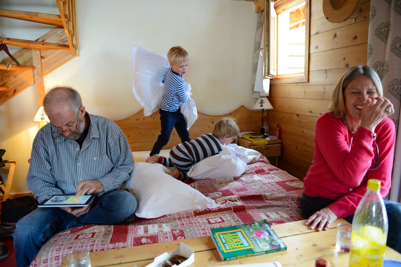 Two children have a playful pillow fight on a bed while two adults sit nearby, one laughing.