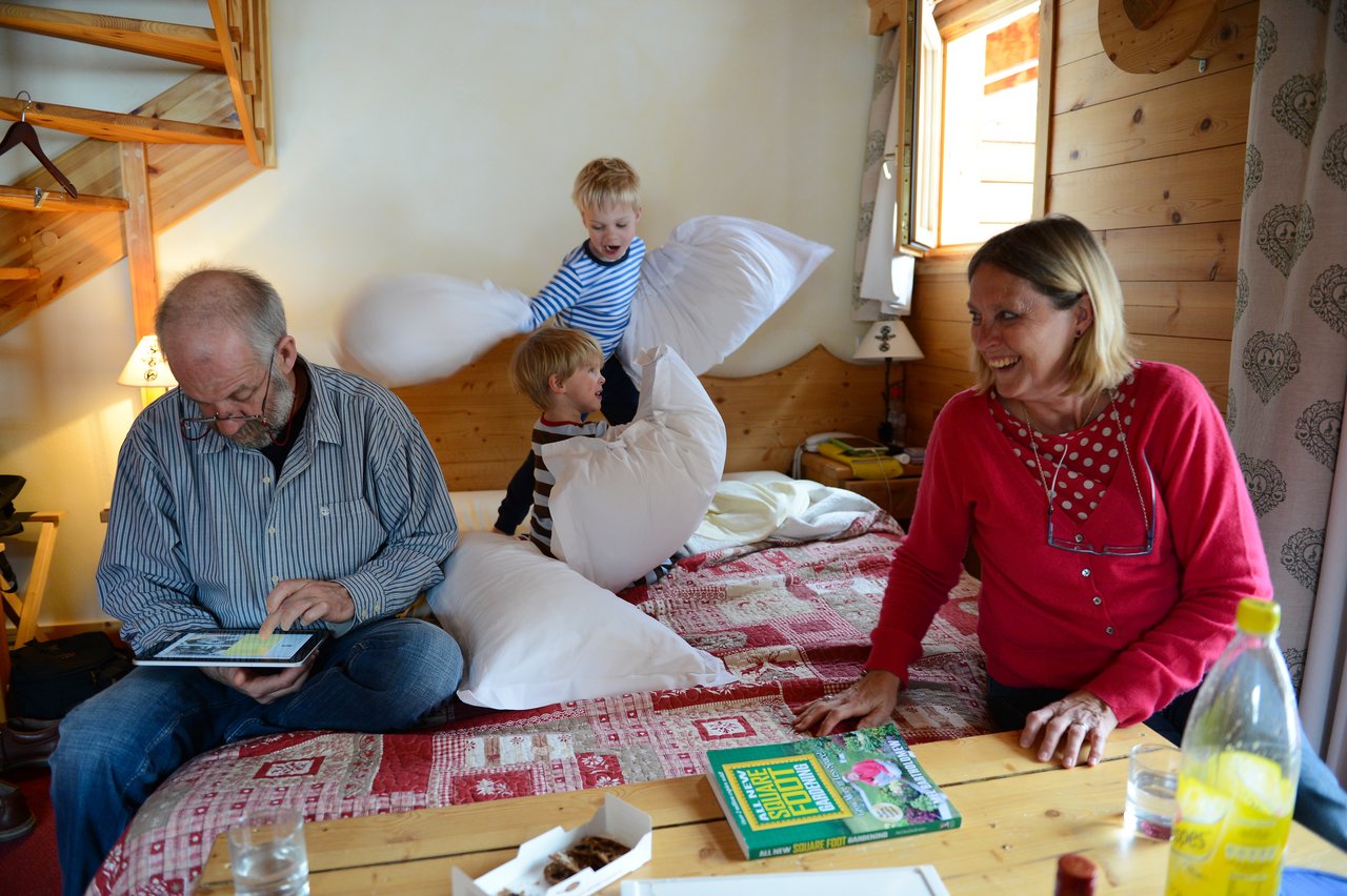 Two children have a playful pillow fight on a bed while two adults sit nearby, one smiling and one reading.