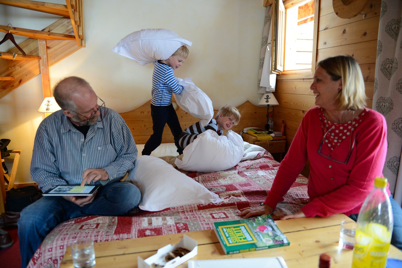 A child swings a pillow at another child on a bed while two adults watch and smile.