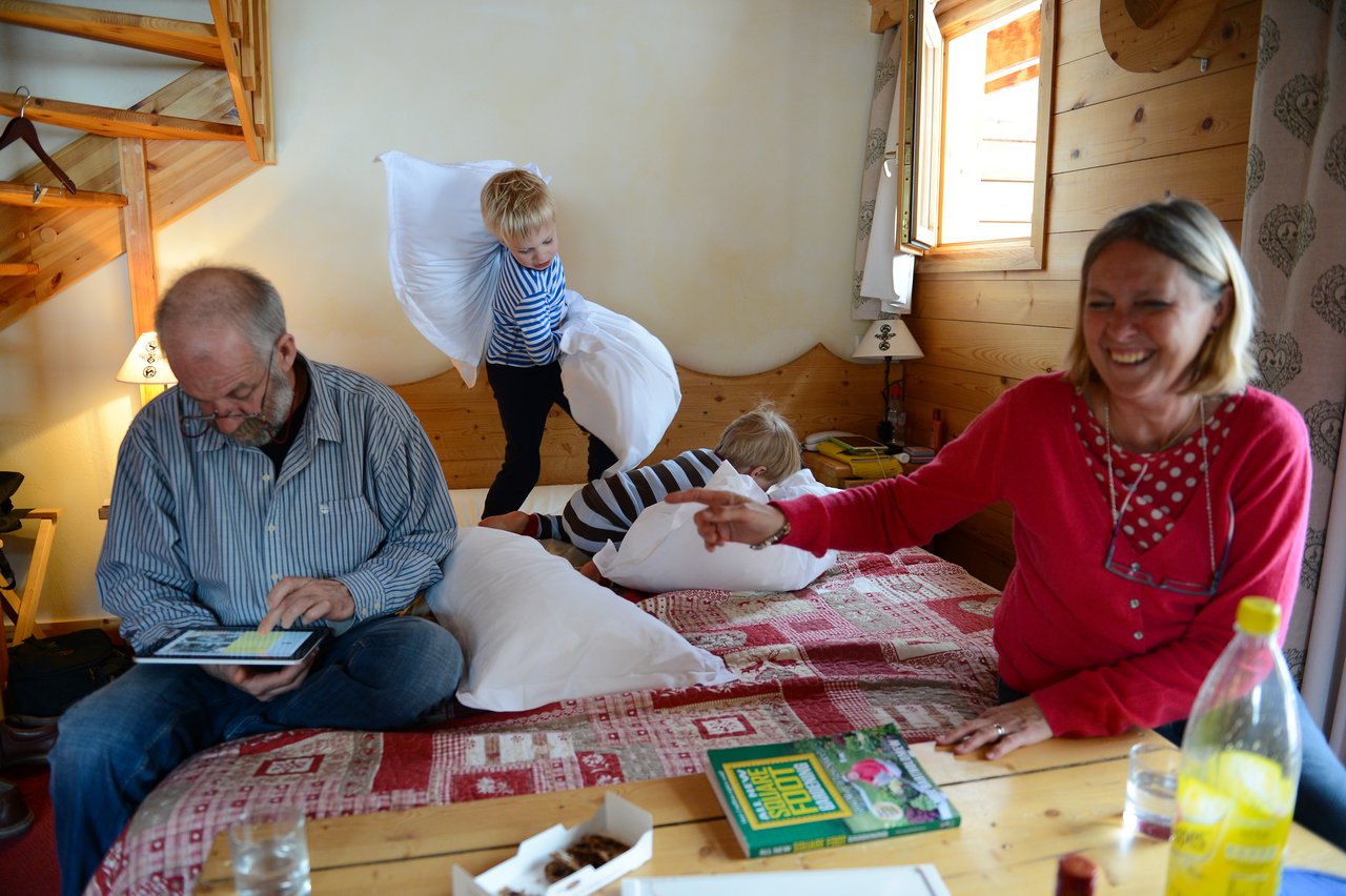 A child swings a pillow during a playful pillow fight on a bed, while others laugh and watch.