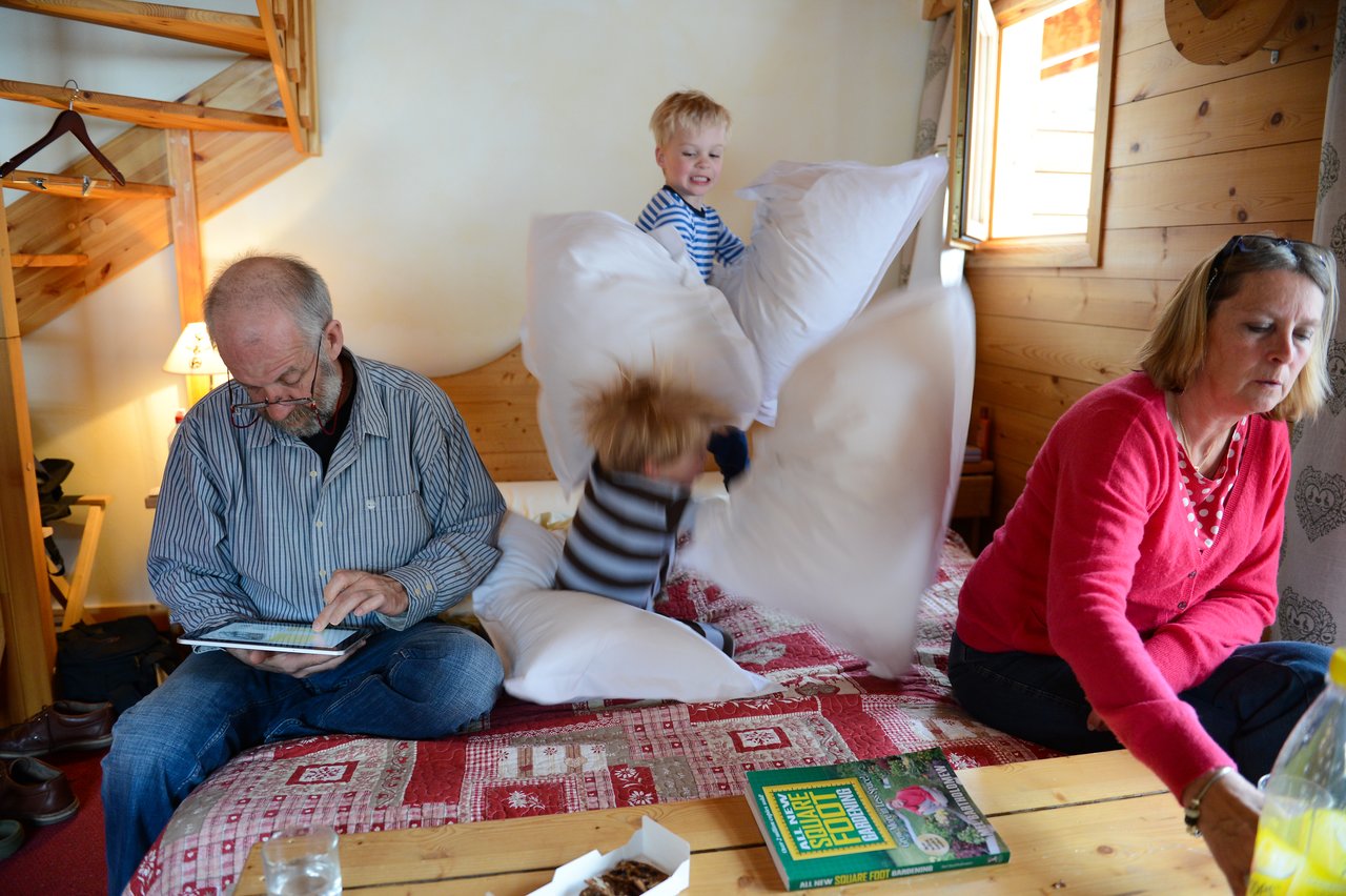 Two children on a bed have a playful pillow fight while two adults sit nearby, focused on other activities.