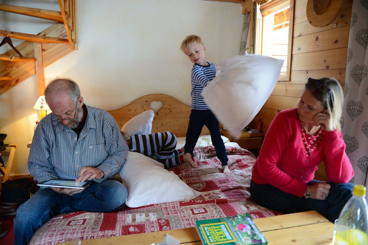 A child playfully swings a pillow on a bed while two adults sit nearby, engaged in other activities.