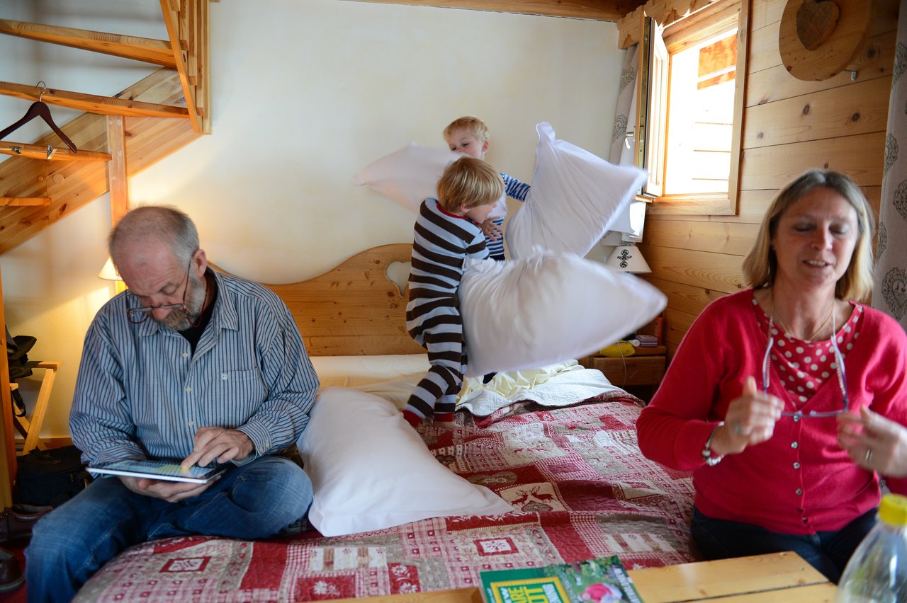 Two children on a bed have a playful pillow fight while two adults sit nearby, engaged in other activities.