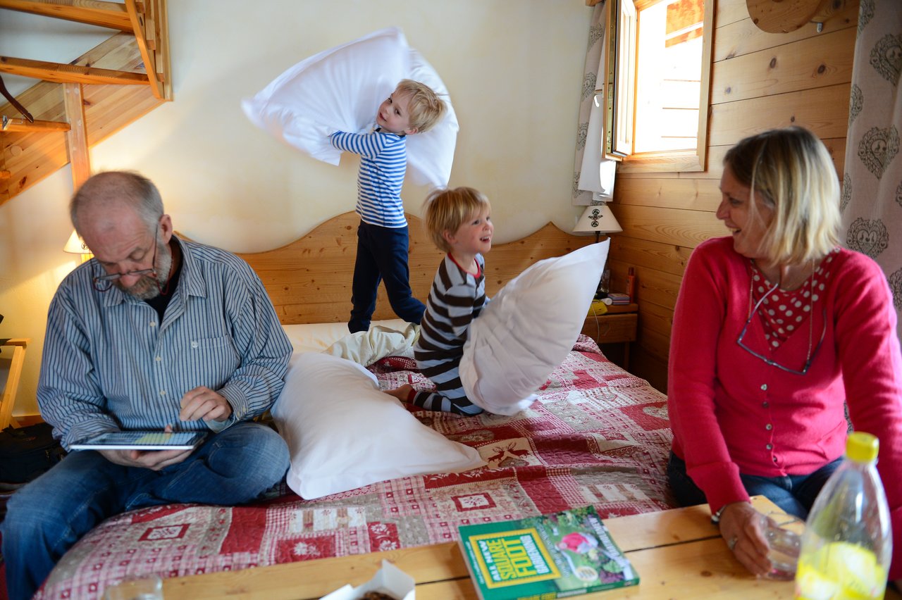 Two children on a bed have a playful pillow fight while two adults sit nearby, one watching and smiling.