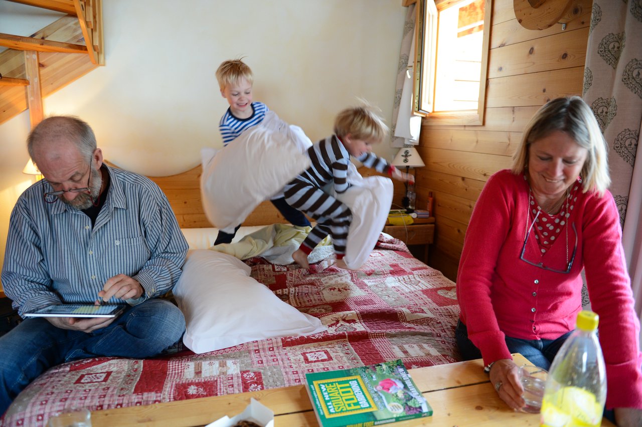 Two children in striped pajamas have a pillow fight on a bed while two adults sit nearby.