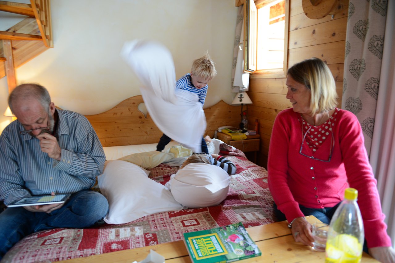 A child energetically swings a pillow on a bed while two adults sit nearby, one watching and smiling.