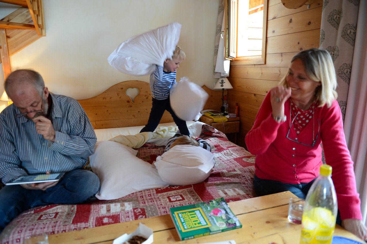 A child energetically swings pillows on a bed while two adults nearby react with amusement and focus on other activities.