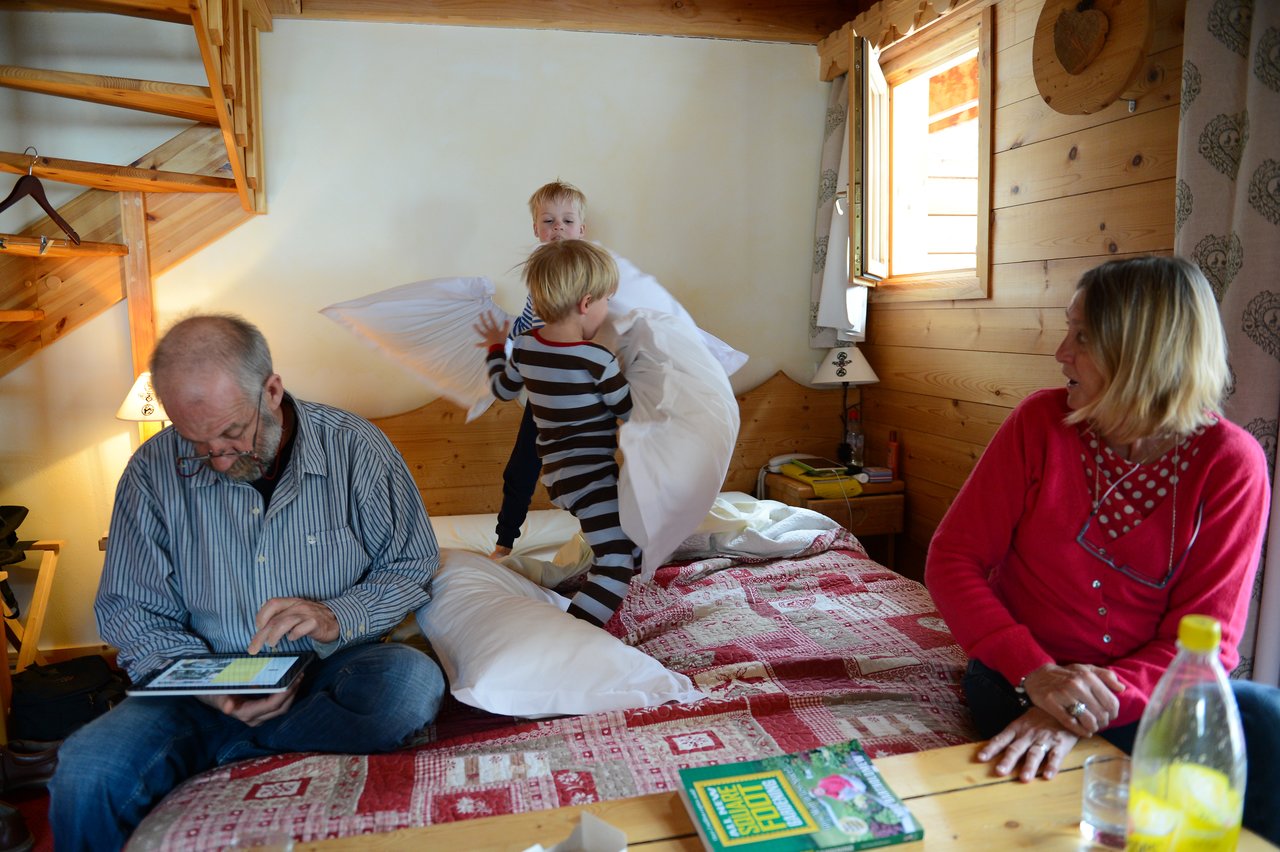 Two children have a playful pillow fight on a bed while two adults watch and another uses a tablet.
