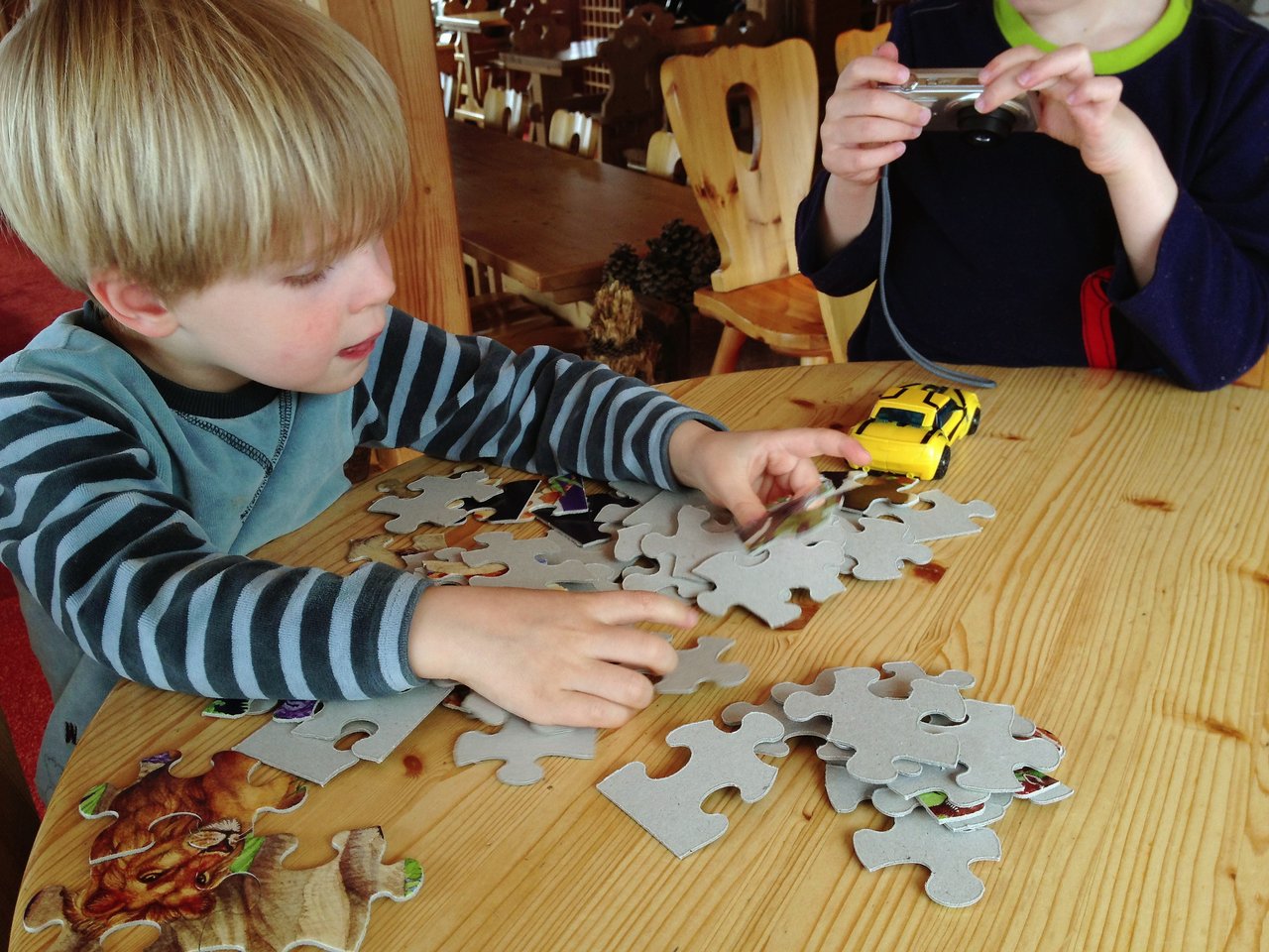 A young child assembles a jigsaw puzzle on a wooden table, while another child holds a small camera.